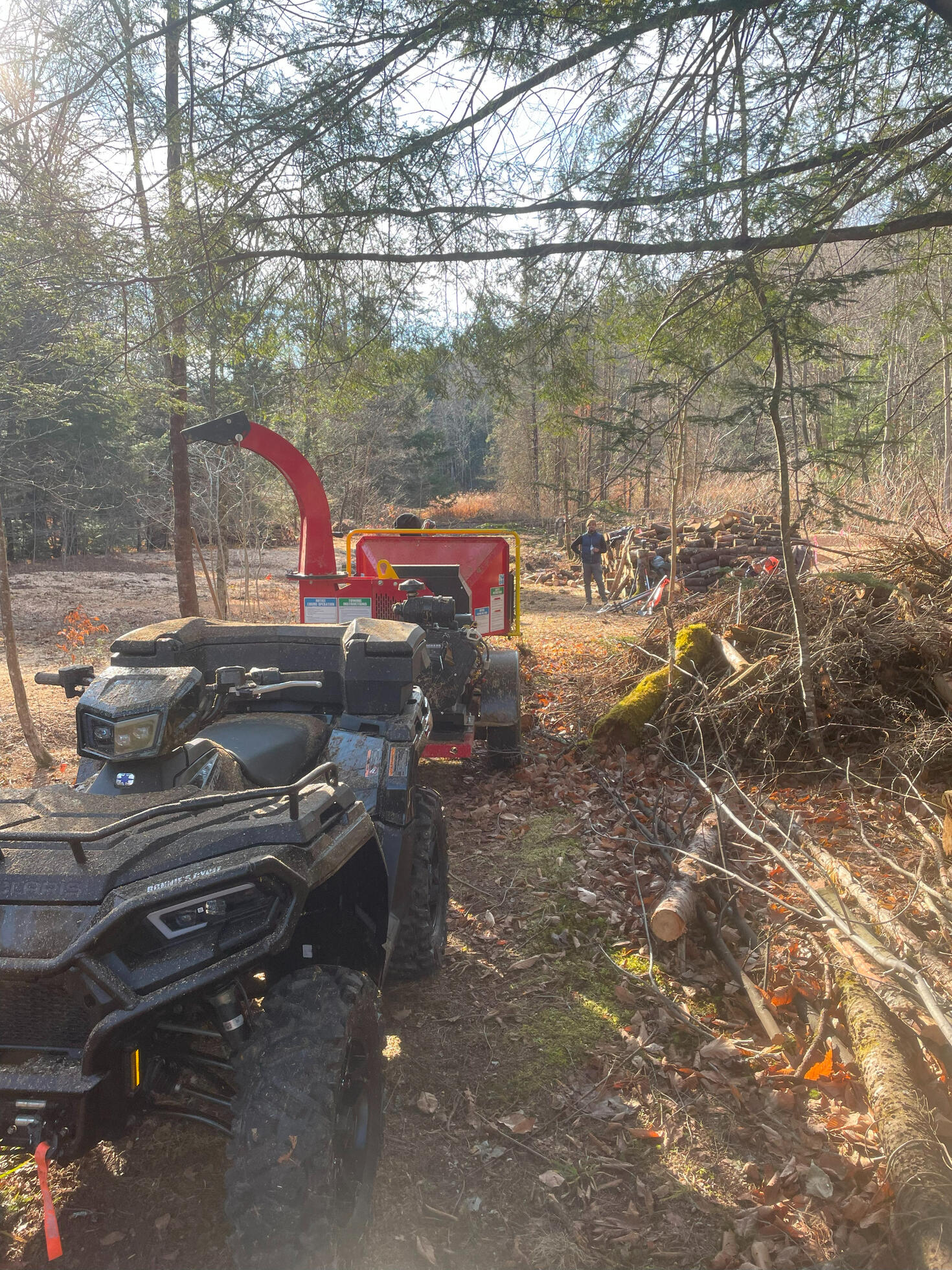 An all-terrain vehicle towing a wood chipper with a person and tools in a forest